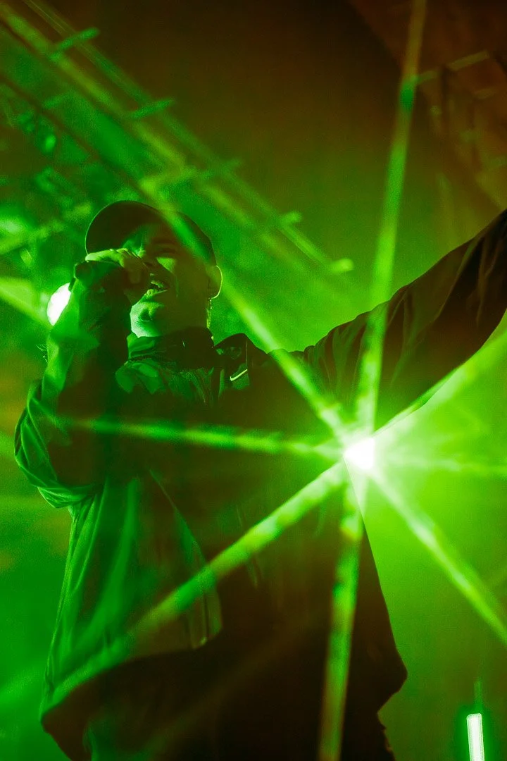 Performer singing or speaking into a microphone on stage, illuminated by green laser lights.
