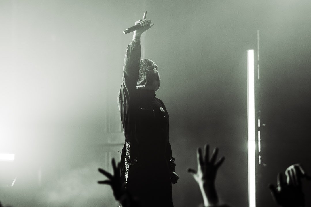 Performer in a hoodie holding a microphone on stage, with hands raised in front, in a black-and-white concert scene.