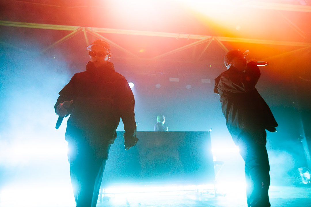 Kneecap performing with hooded jackets and sunglasses on stage, holding microphones, with DJ Próvaí in the background, colourful stage lighting, and fog effects.