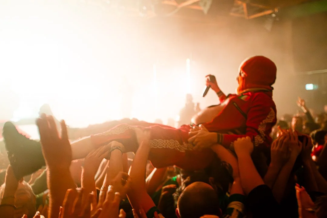 A concert crowd crowdsurfs at a lively music event with bright stage lights in the background.