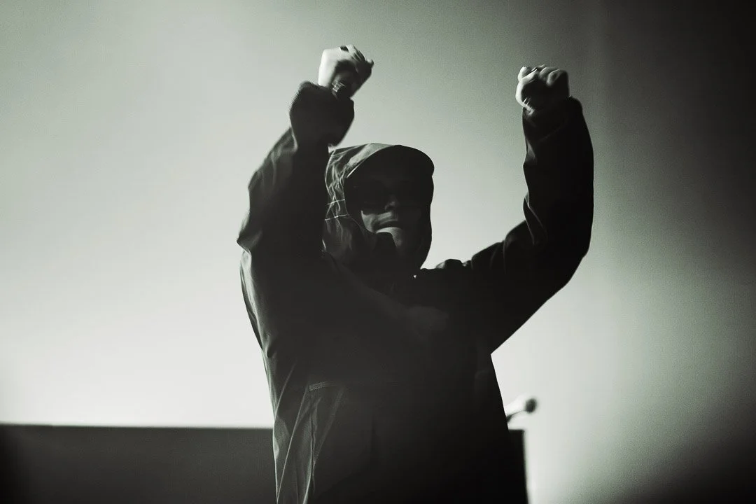 A person wearing a hooded jacket and sunglasses raising both fists in the air, silhouette in front of a plain background.