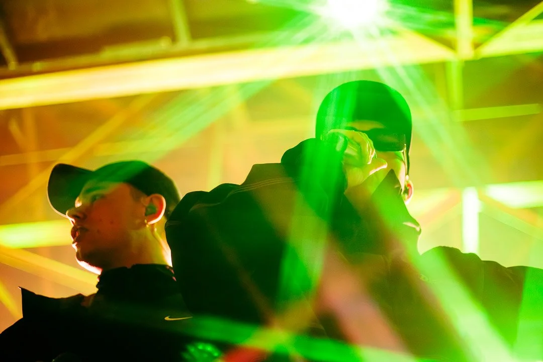 Two men performing on stage with green and yellow lights shining around them, one is holding a microphone and the other has a cap and glasses.