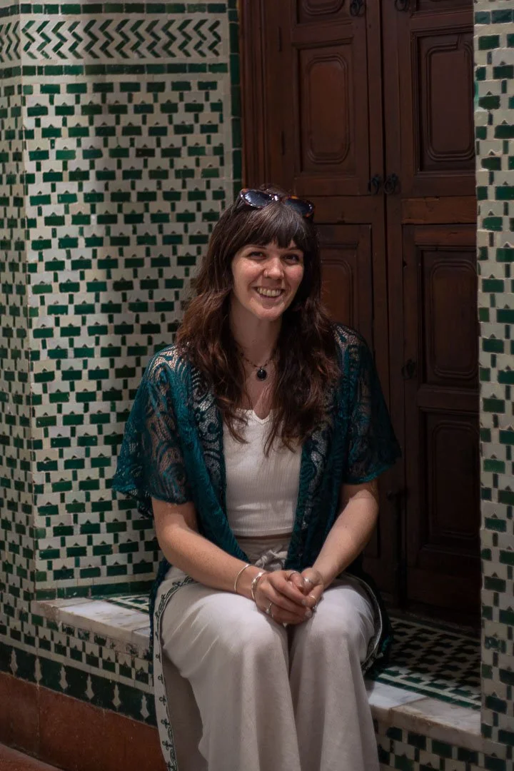 A woman with brown hair and bangs, wearing a white top, a blue lace shawl, and white pants, sitting on a tiled ledge in front of a green and white tiled wall with a wooden cabinet behind her, smiling at the camera.