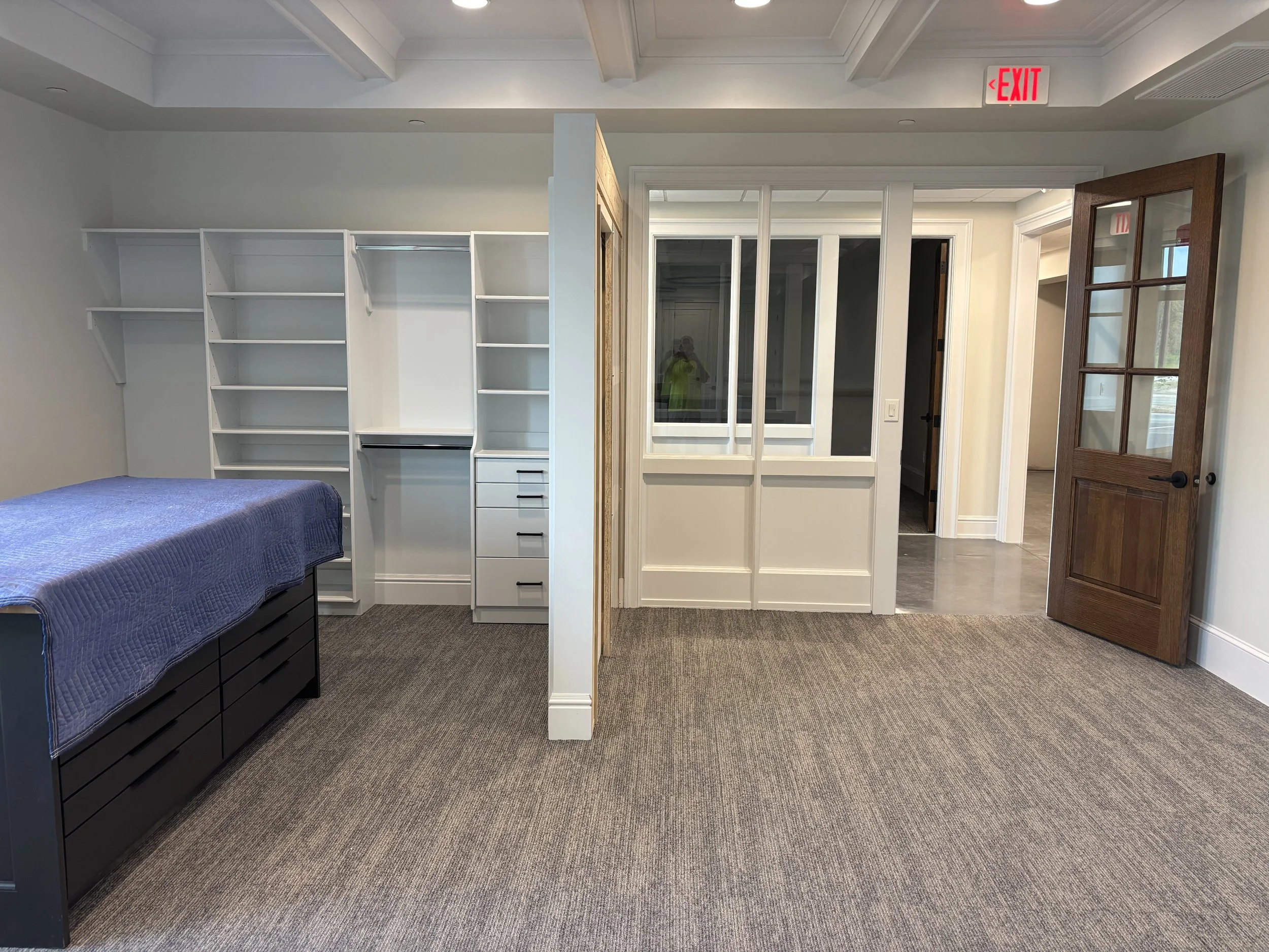 Empty room with built-in white shelves, a bed with a blue cover, and glass-paneled doors leading to other areas, with an exit sign overhead.