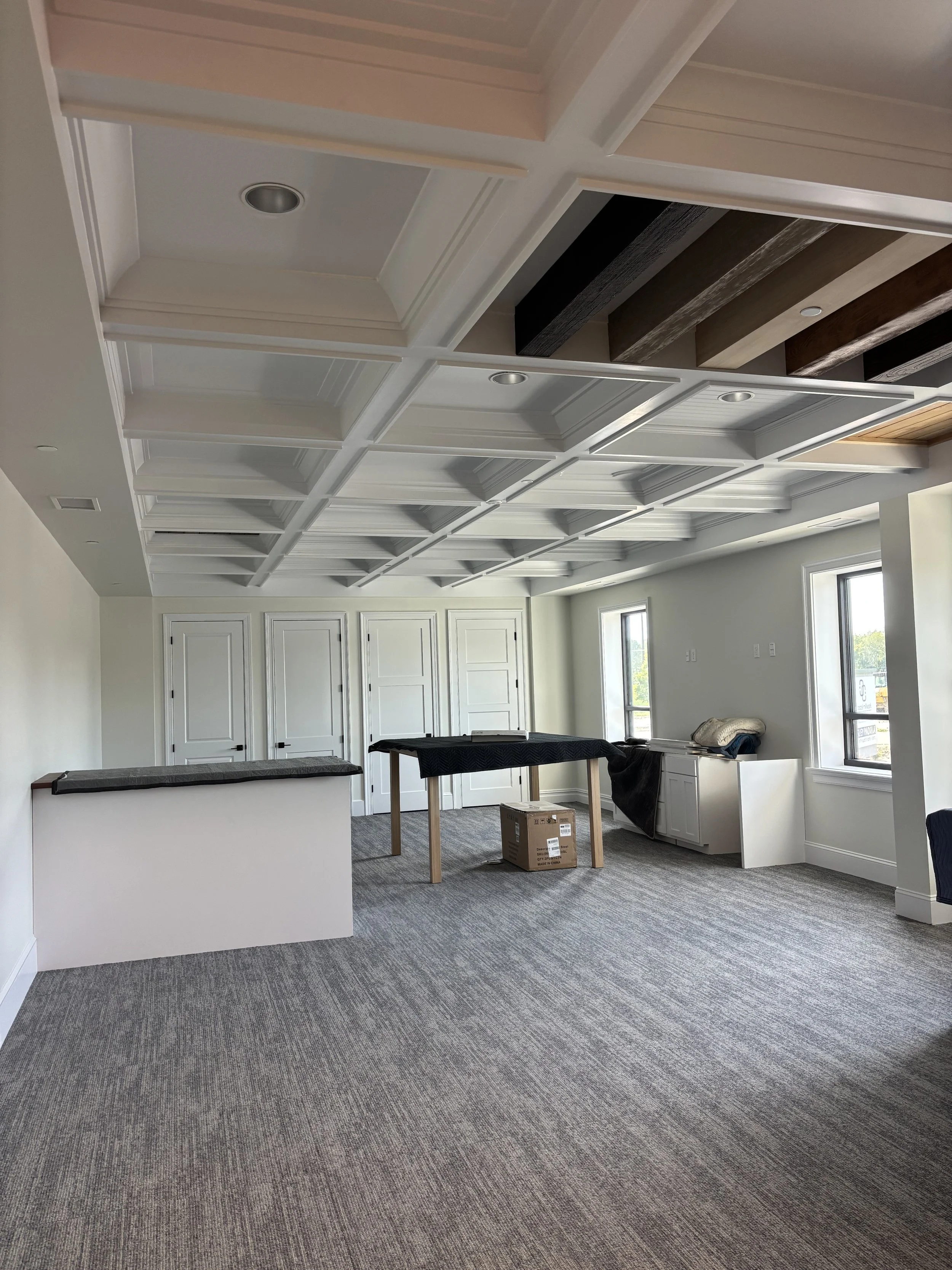 Empty room with decorative coffered ceiling, carpeted floor, two windows, white walls, and white doors in the background. There are some furniture and boxes arranged in the room.