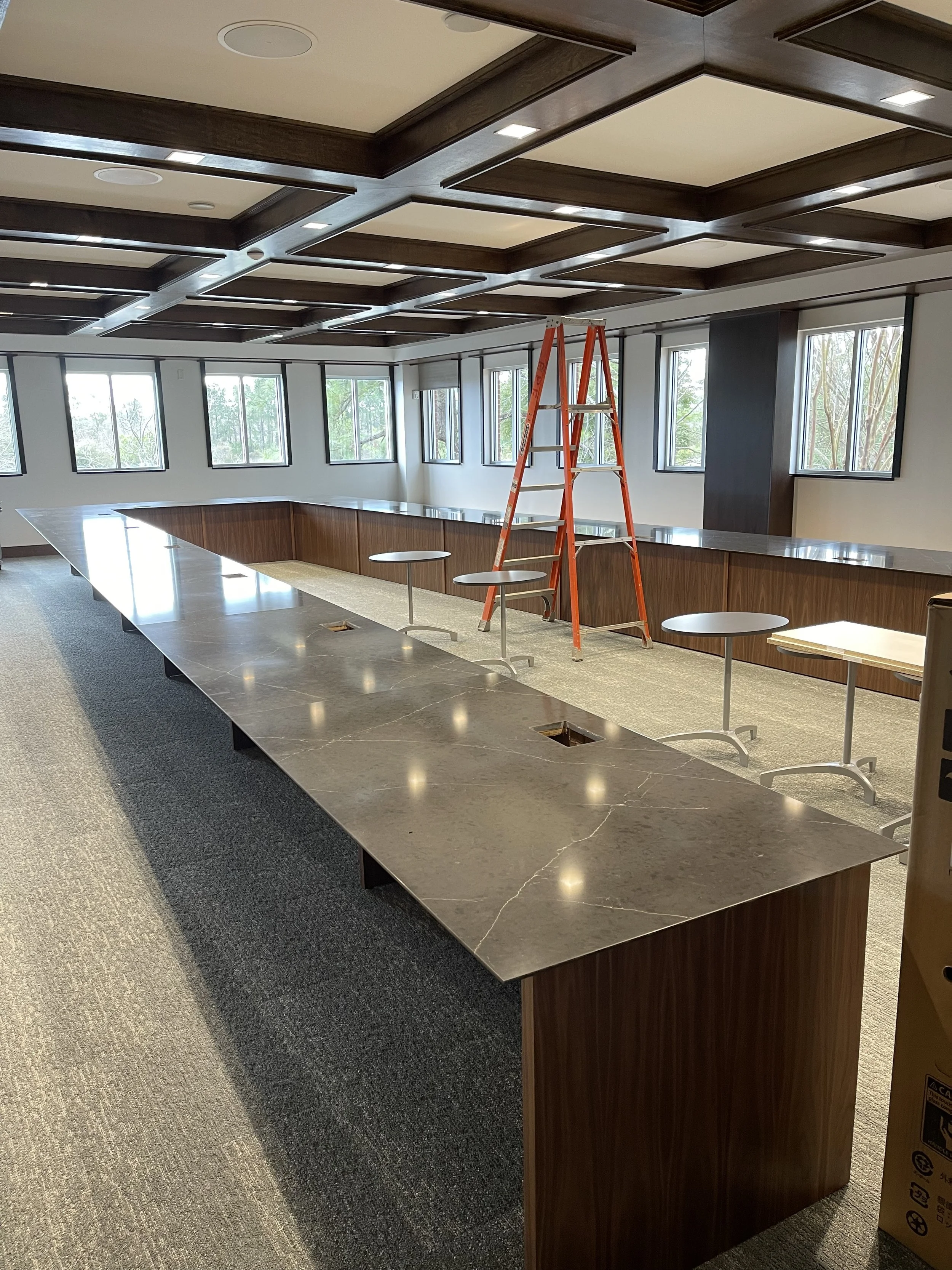 Empty conference room with marble-topped table, a ladder, and small round tables, with large windows and a coffered ceiling.