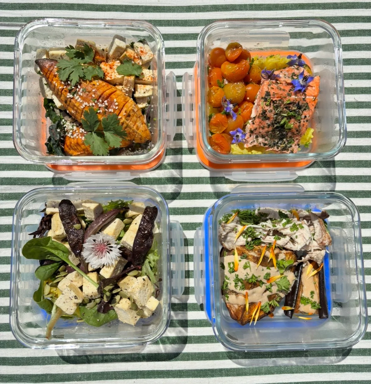 Four glass containers with plated food on a green and white striped tablecloth. Top left: grilled vegetables and tofu garnished with cilantro and sesame seeds. Top right: roasted cherry tomatoes and a piece of salmon with herbs and edible flowers. Bottom left: mixed salad with tofu, greens, and edible flowers. Bottom right: cooked fish fillet garnished with herbs and sliced peppers.