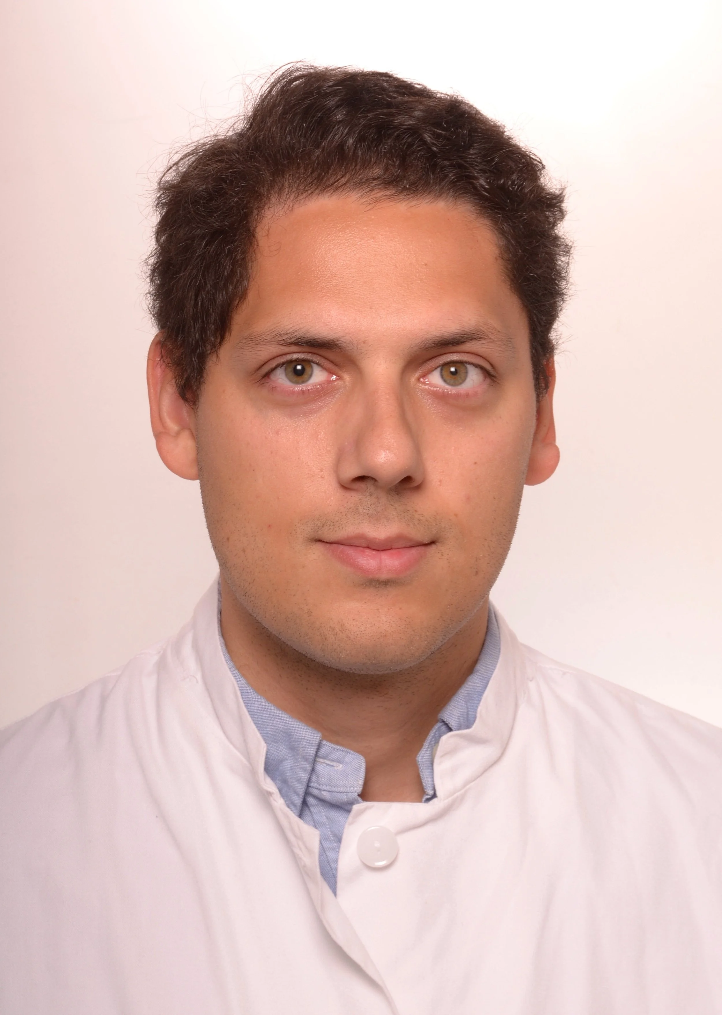 Portrait of a young man with brown hair, green eyes, and light skin, wearing a white chef's jacket with a blue collared shirt underneath, standing against a plain white background.
