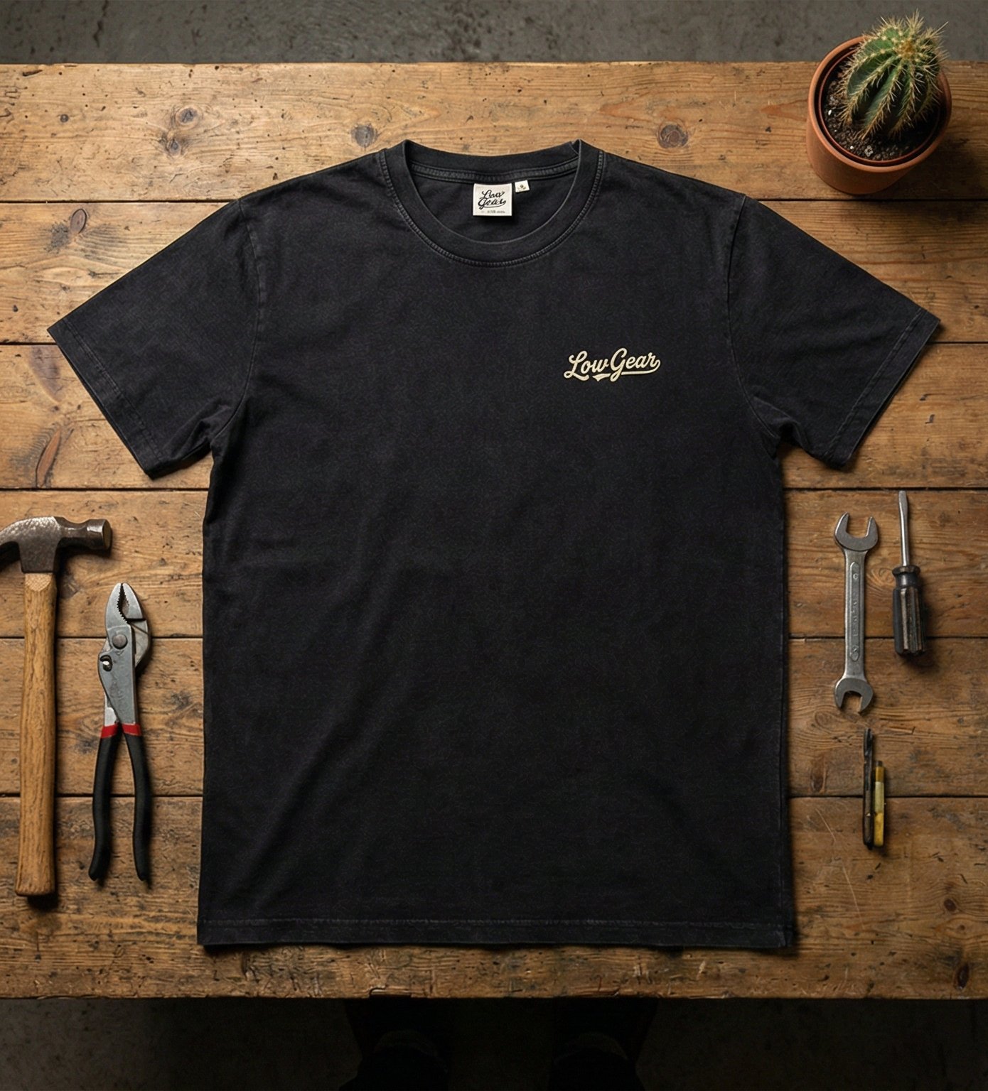 A black T-shirt with the words "Low Gear" printed on the chest, laid out on a wooden workbench surrounded by tools and a small potted cactus.