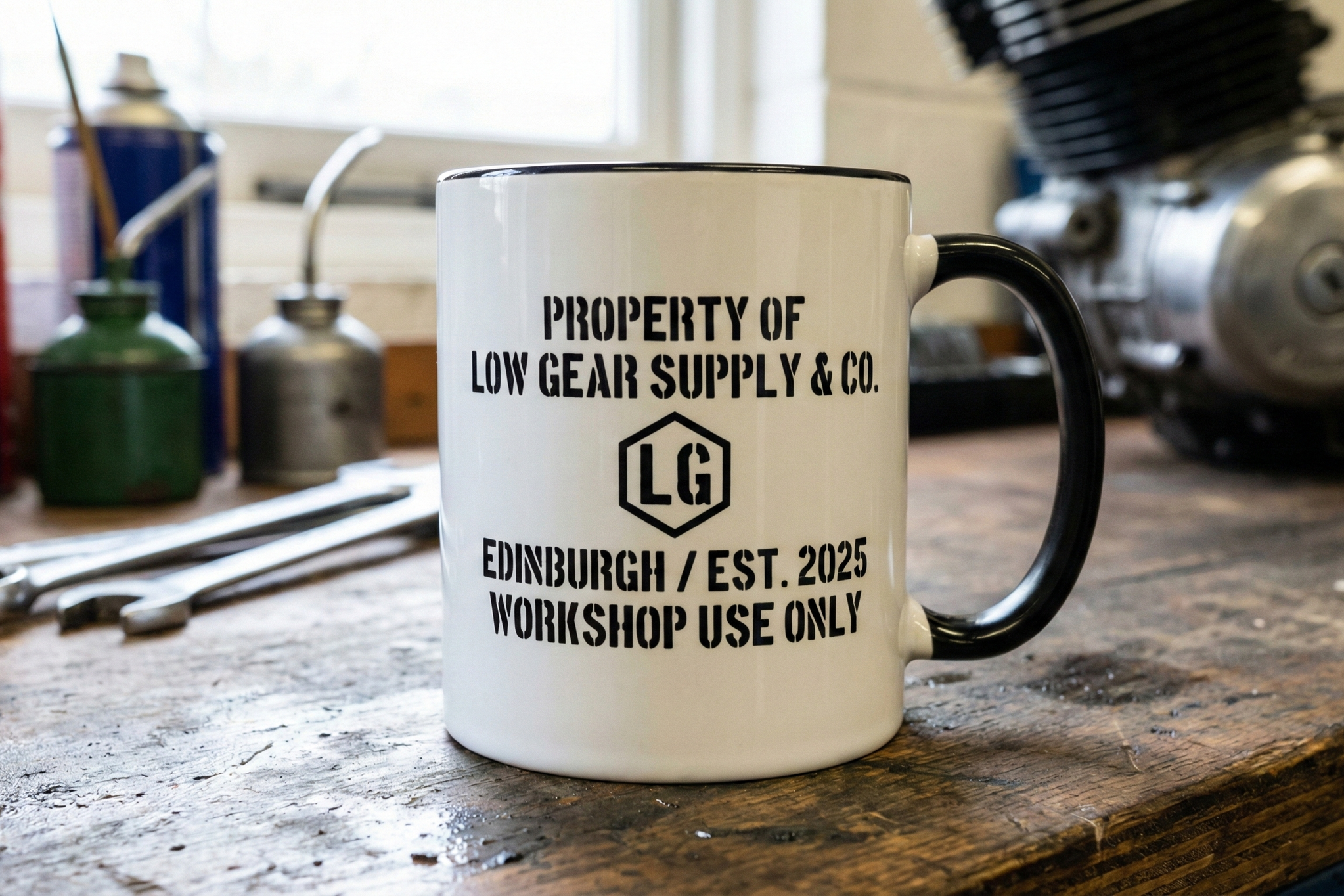 A coffee mug with the logo and text of Low Gear Supply & Co., on a wooden workbench with tools and workshop equipment in the background.