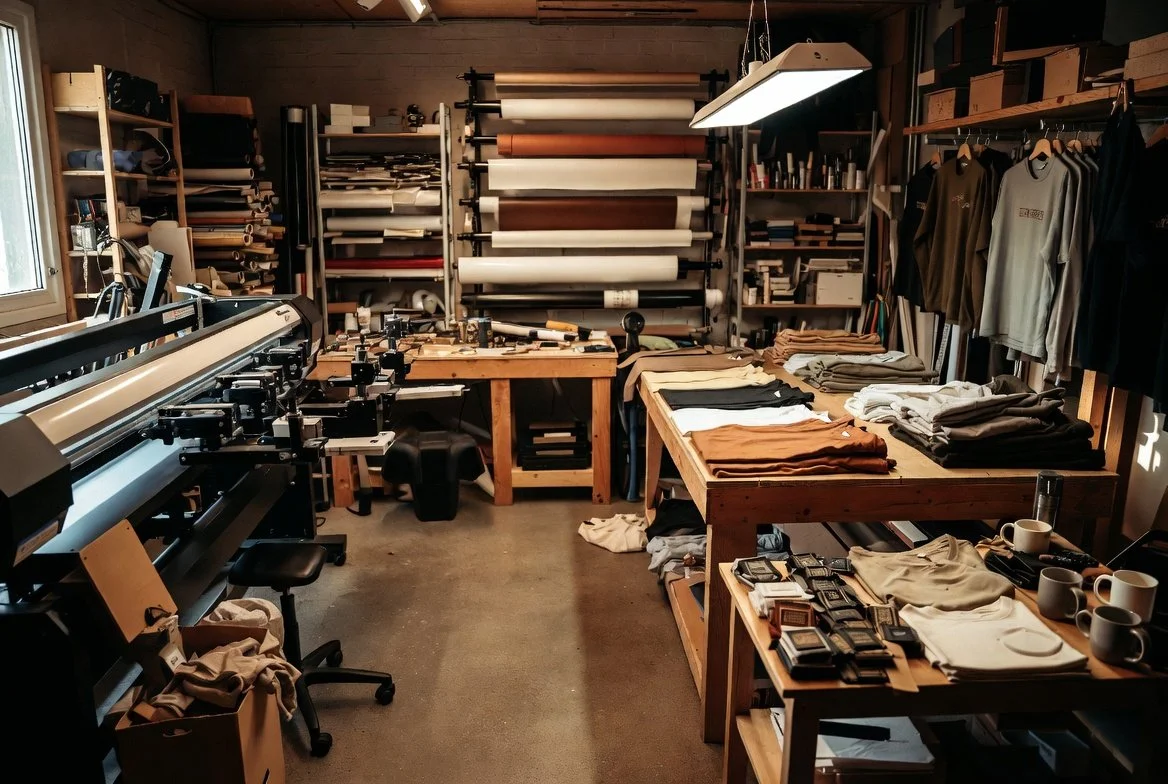 Interior of a custom printing workshop with large format printer, shelves of rolls of paper and fabric, tables with folded clothes, and various printing supplies.