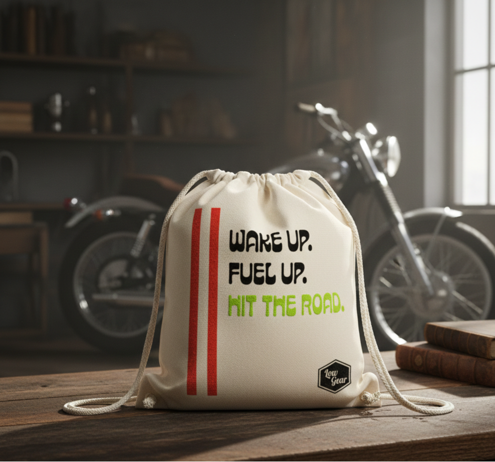 A white drawstring bag with motivational text on it in black, green, and red, placed on a wooden table in a room with a motorcycle in the background and books on the side.