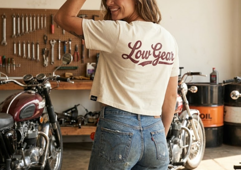 A smiling woman in a white crop top with 'Low Gear' text on the back, standing in a garage next to motorcycles and tools.