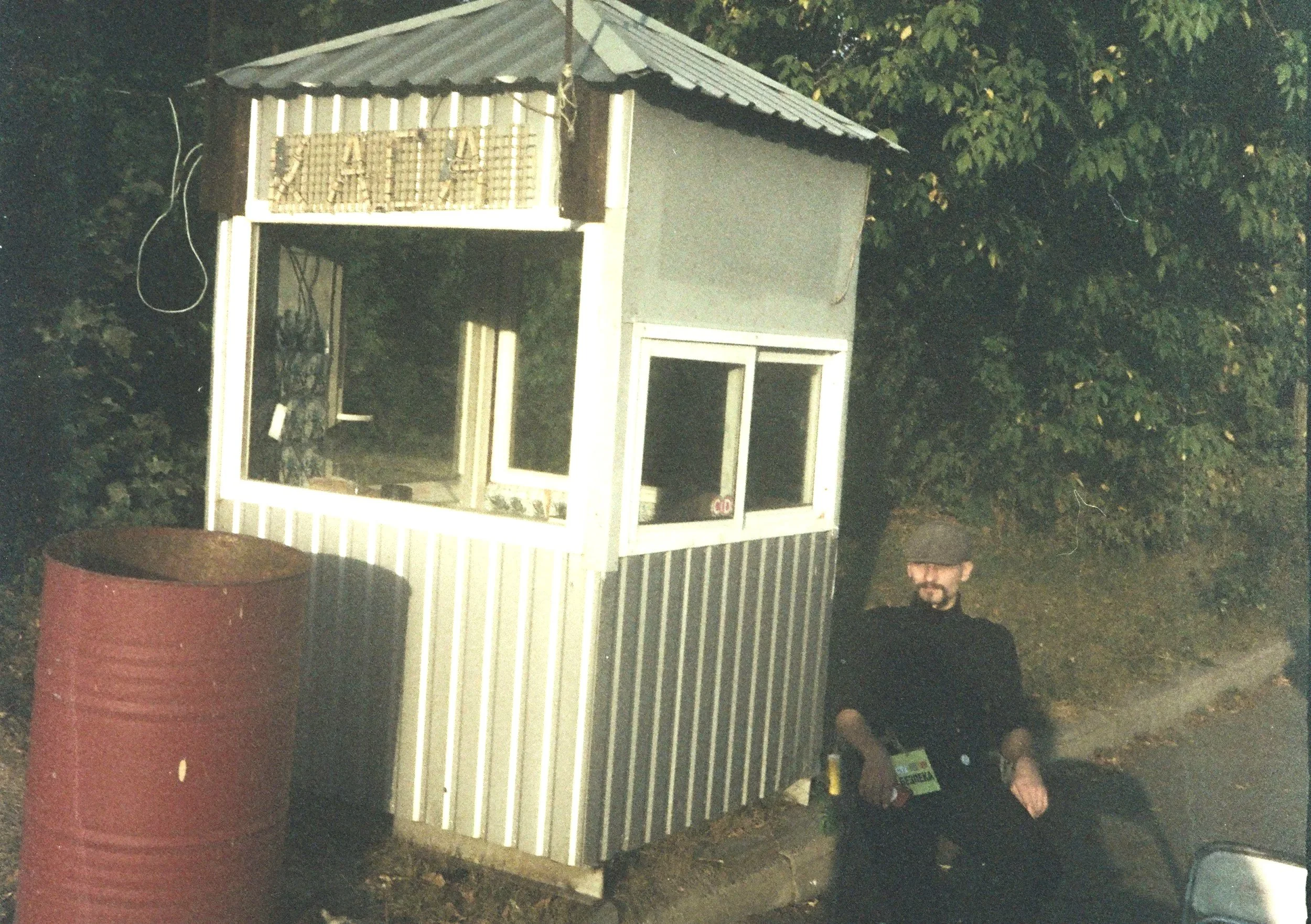 A man wearing a hat and glasses sitting beside a small roadside retail stand or kiosk with striped siding and glass windows, next to a large rusty barrel, with trees in the background.