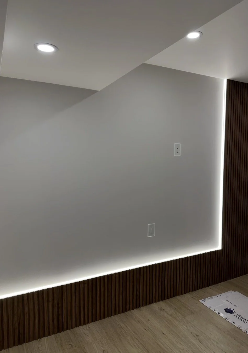 Modern interior wall with recessed lighting, electrical outlets, a wooden baseboard, and a vertical wooden panel on the right side, with a strip of LED lighting along the baseboard.
