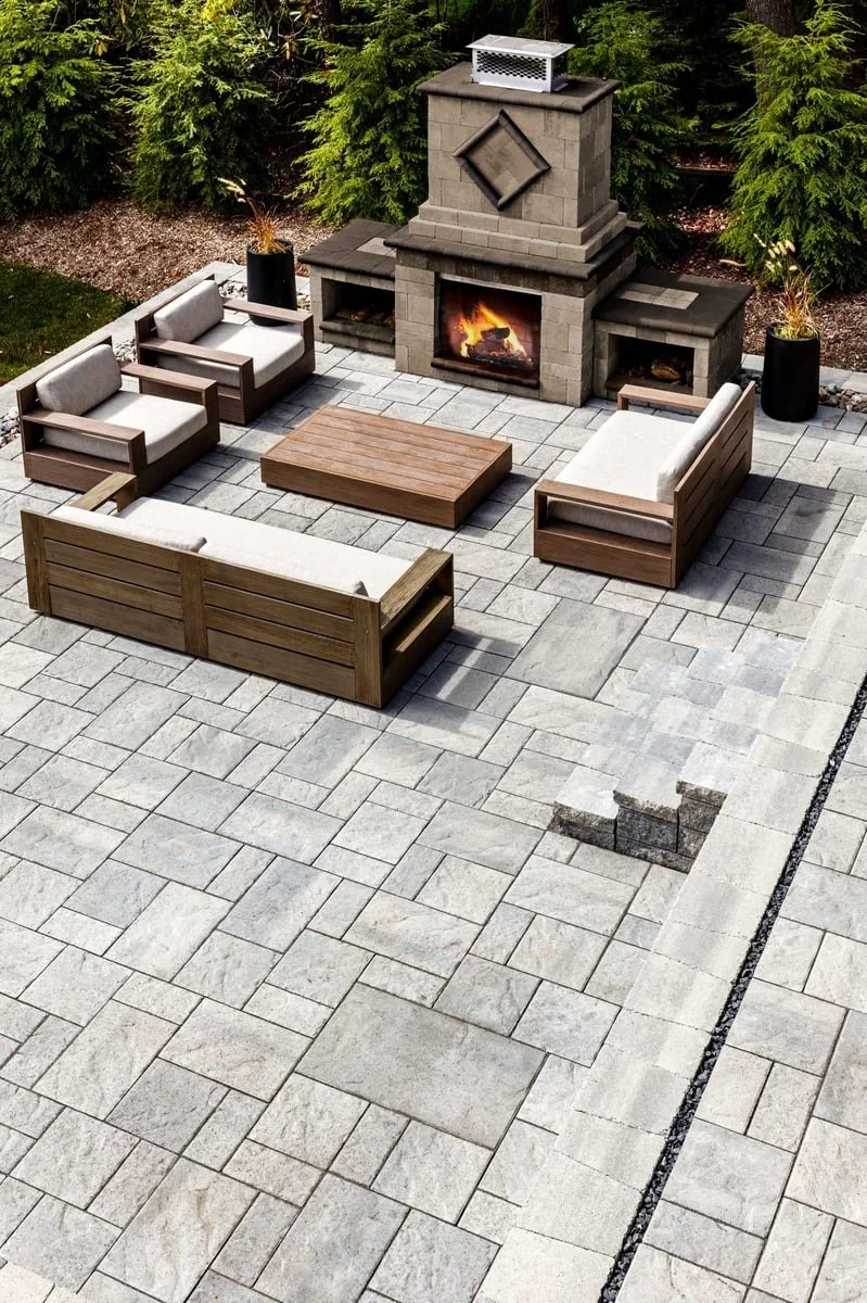 Outdoor patio with a stone fireplace, surrounded by outdoor furniture including cushioned sofas and chairs, and potted plants, on a paved stone surface.