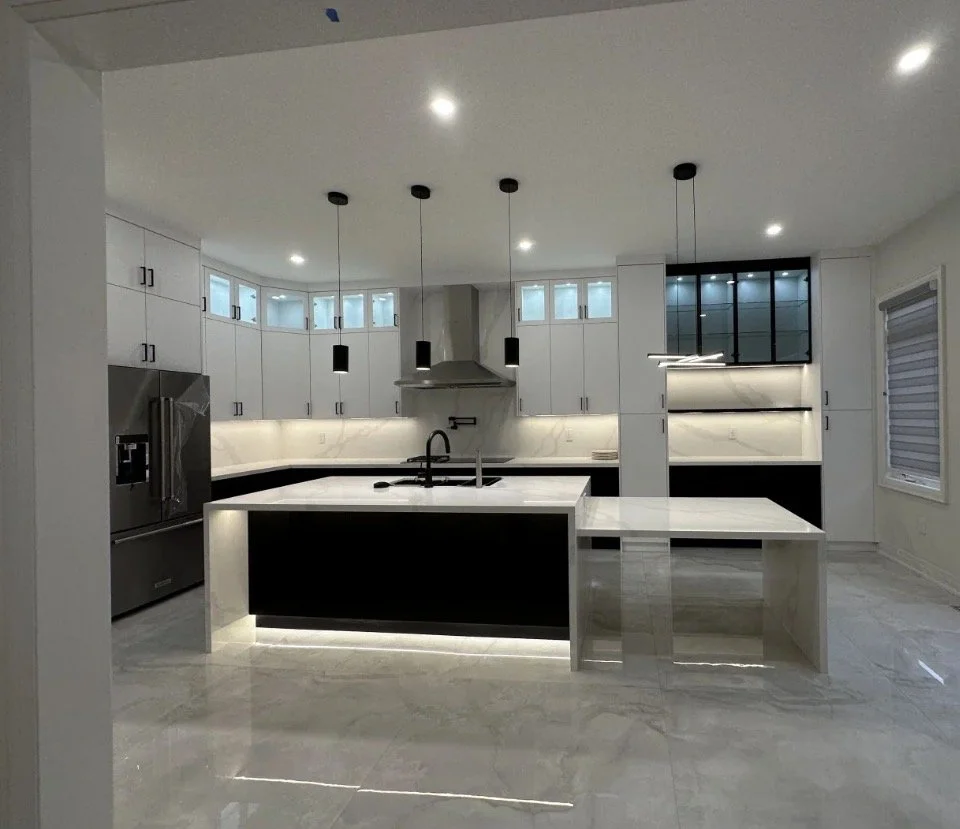 Modern kitchen with white cabinets, black countertops, central island with black panel, stainless steel refrigerator, and built-in oven. Pendant lights hang above the island.