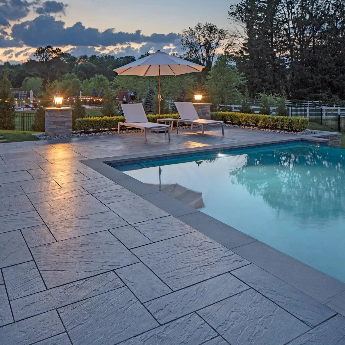 An outdoor swimming pool surrounded by a stone patio with two lounge chairs and a white patio umbrella, lit by small lamps, with trees and a fence in the background at dusk.