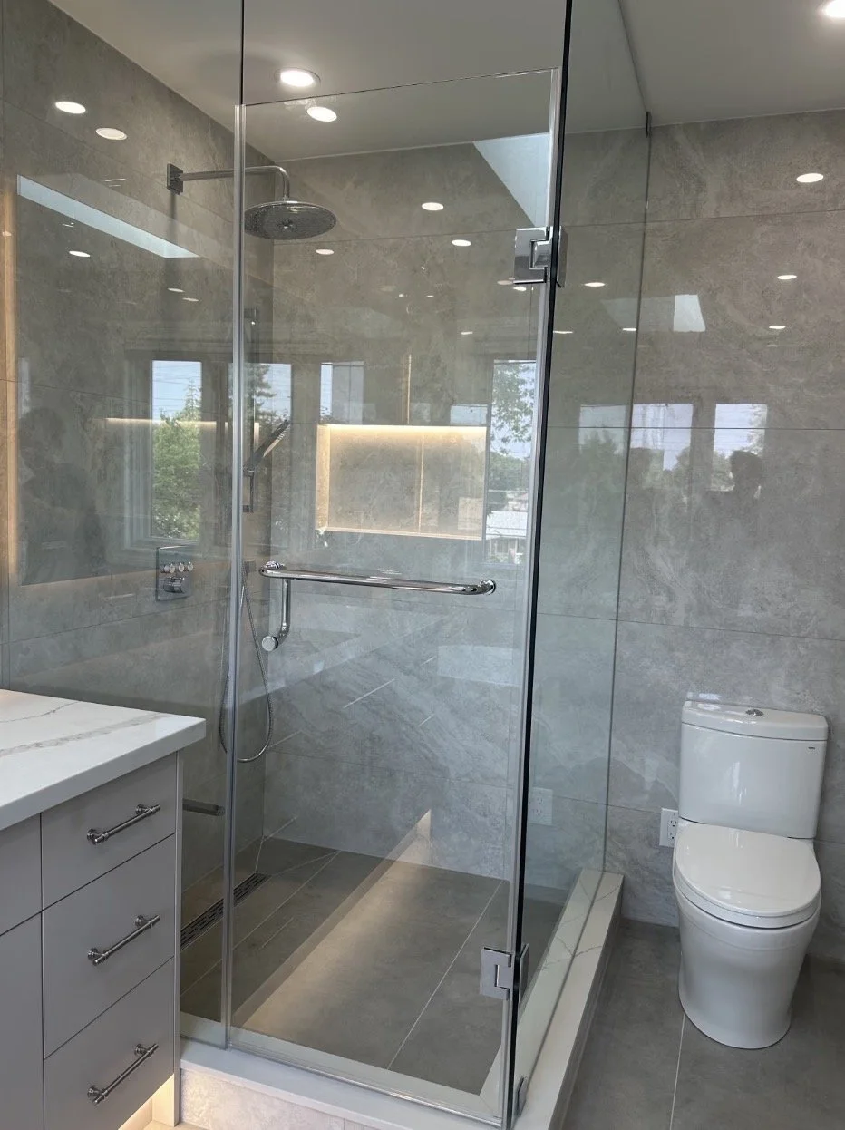 Modern bathroom with a walk-in shower enclosed by glass, a rainfall showerhead, a handheld shower, and gray stone tiles. There is a white toilet to the right and a gray vanity with drawers and a white countertop on the left. Reflections of windows and ceiling lights are visible.