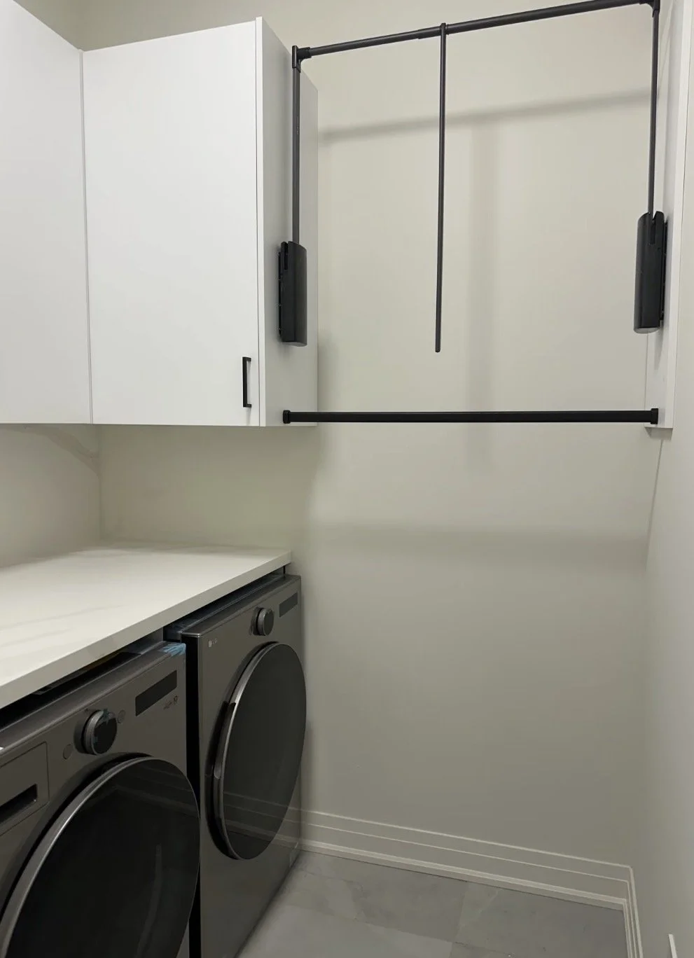A laundry room with a white cabinet, two front-loading washer and dryer, white countertops, and a black pull-up pole attached to the wall for hanging clothes.