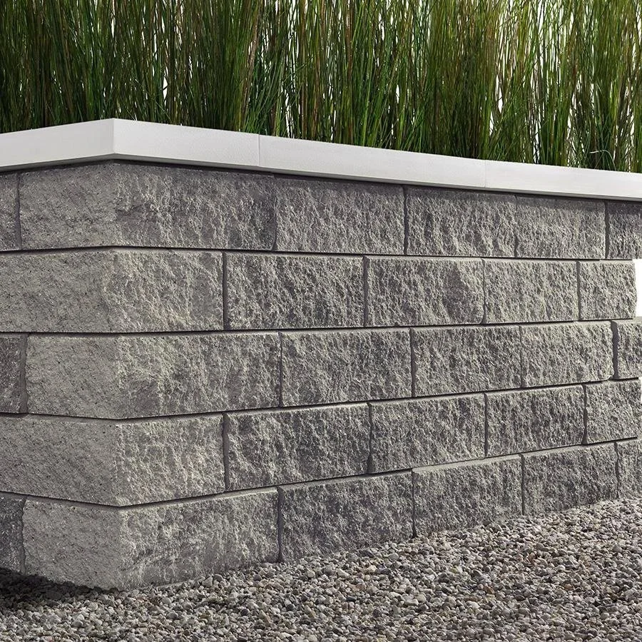 Close-up of a stone block retaining wall topped with a white trim and filled with tall green grasses, with gravel at the base.