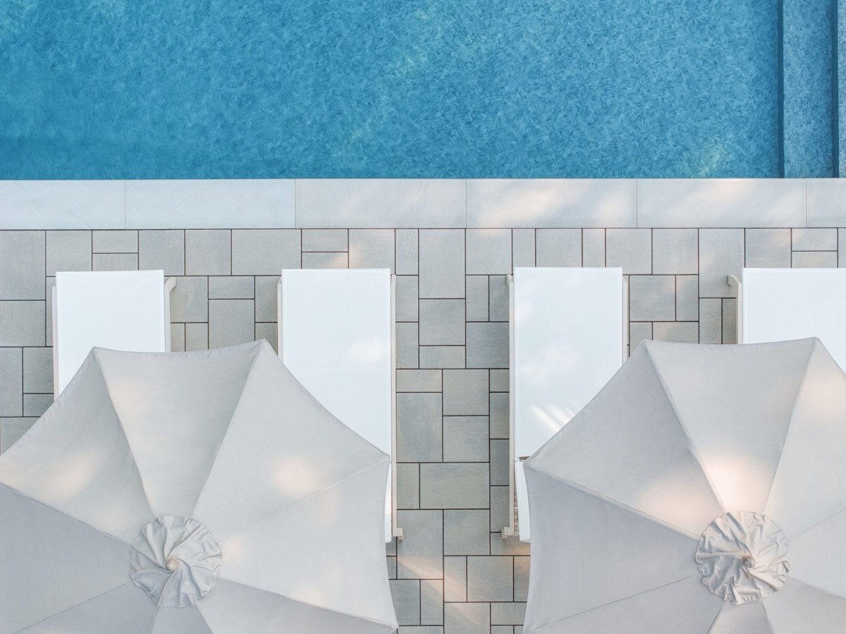 Top view of a swimming pool with four white lounge chairs and two closed white umbrellas on a tiled pool deck.