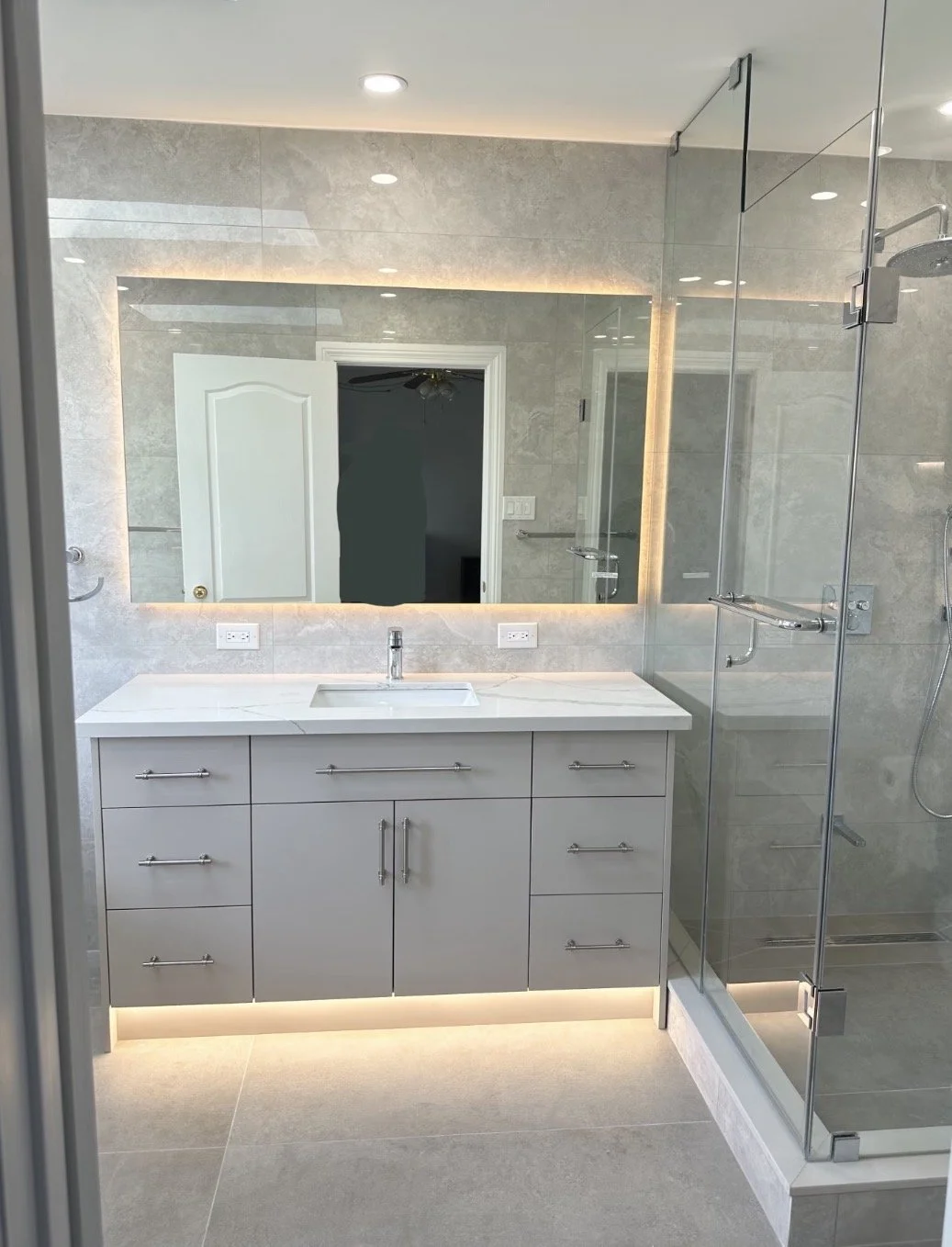 Modern bathroom with large mirror, white vanity with a rectangular sink, gray cabinets, and a glass-enclosed shower with a rainfall showerhead. Recessed lighting and gray tiled walls and floor.