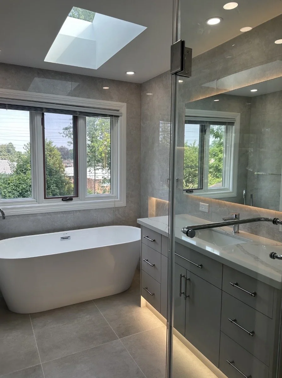 Bathroom with freestanding bathtub, large window with view of greenery, and a vanity with gray cabinets and a large mirror.