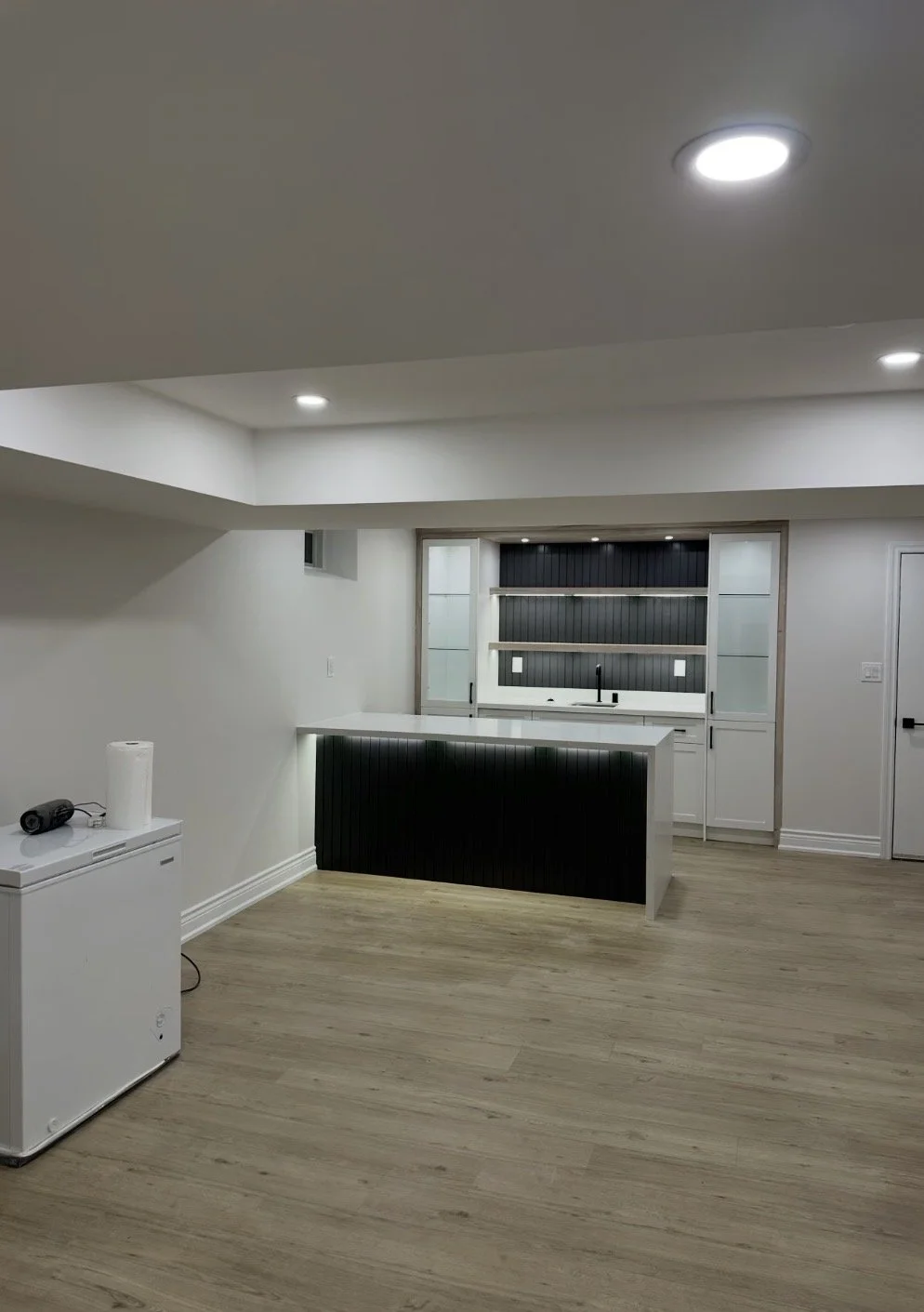 A modern kitchen with white walls, light wood flooring, an island with a white countertop and black paneling, and a windowed area with dark backsplash and white cabinetry.