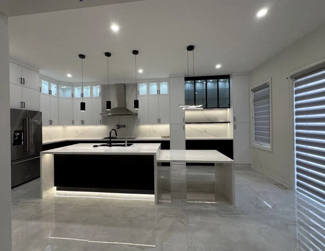 Modern kitchen with white cabinets, black accents, marble countertops, and pendant lights, featuring a large kitchen island with a sink, stainless steel refrigerator, and a patterned floor.