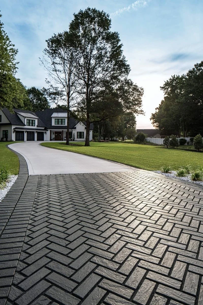 Residential driveway with herringbone brick pavers in the foreground, leading to a concrete driveway and modern house with black roofs and white walls, surrounded by green grass and trees, during dusk.
