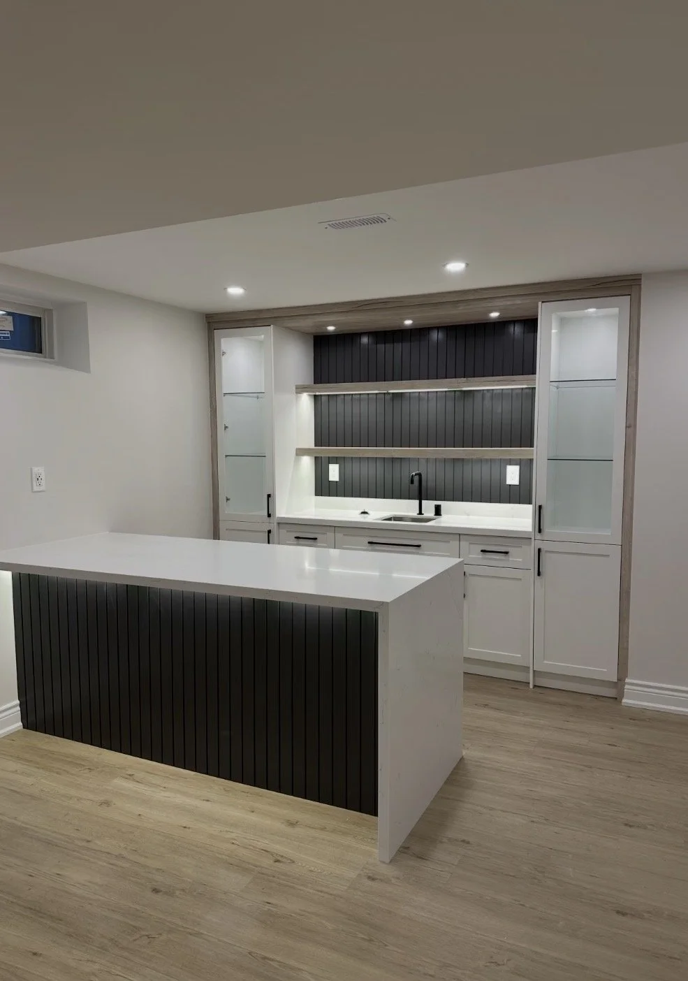 Modern kitchen with white cabinets, a black backsplash, and an island with black paneling. Recessed lighting and wooden flooring.