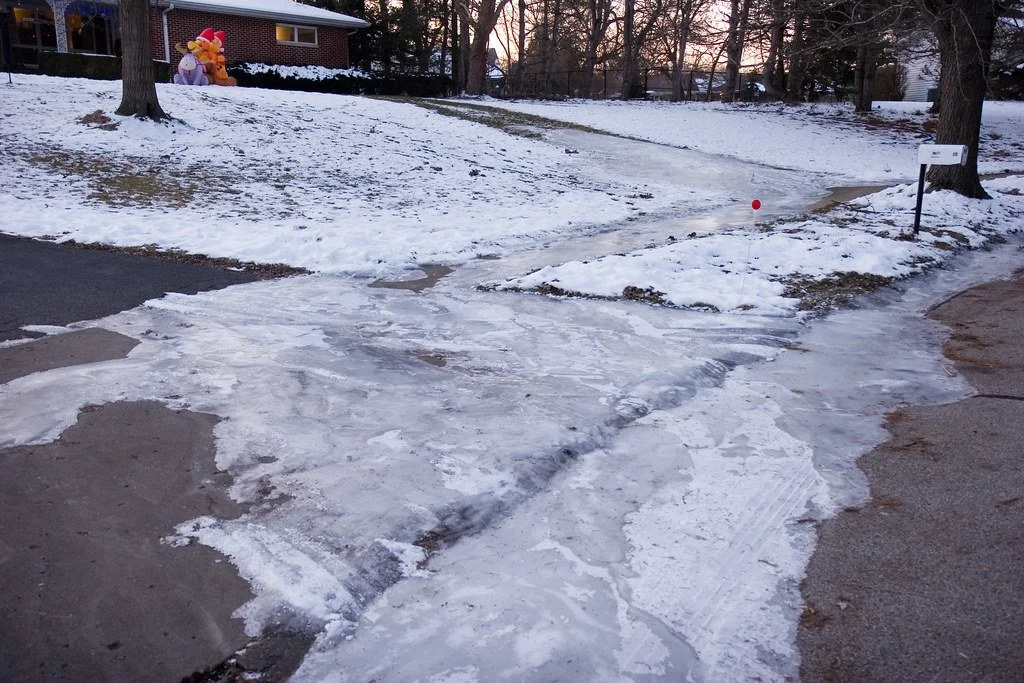 A snow-covered sidewalk with patches of ice, a red ball, a small mailbox, a home in the background, and ice sculptures of a giraffe and a lion on the lawn.