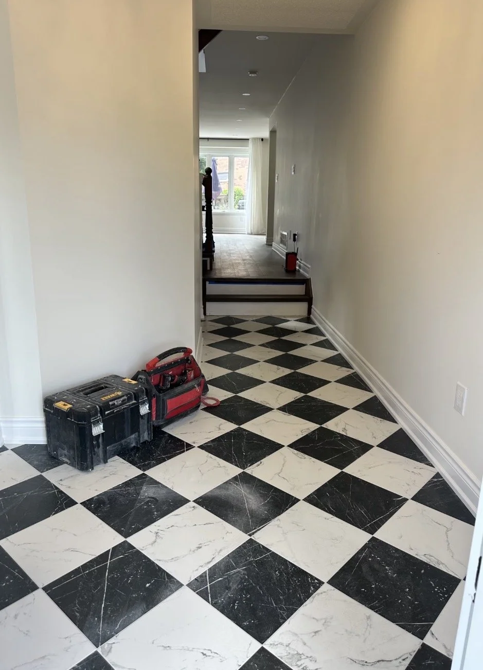 An entryway with black and white checkered marble flooring, some tool boxes and a red bag on the floor, leading to a staircase and a sunlit living room with a large window and curtain.