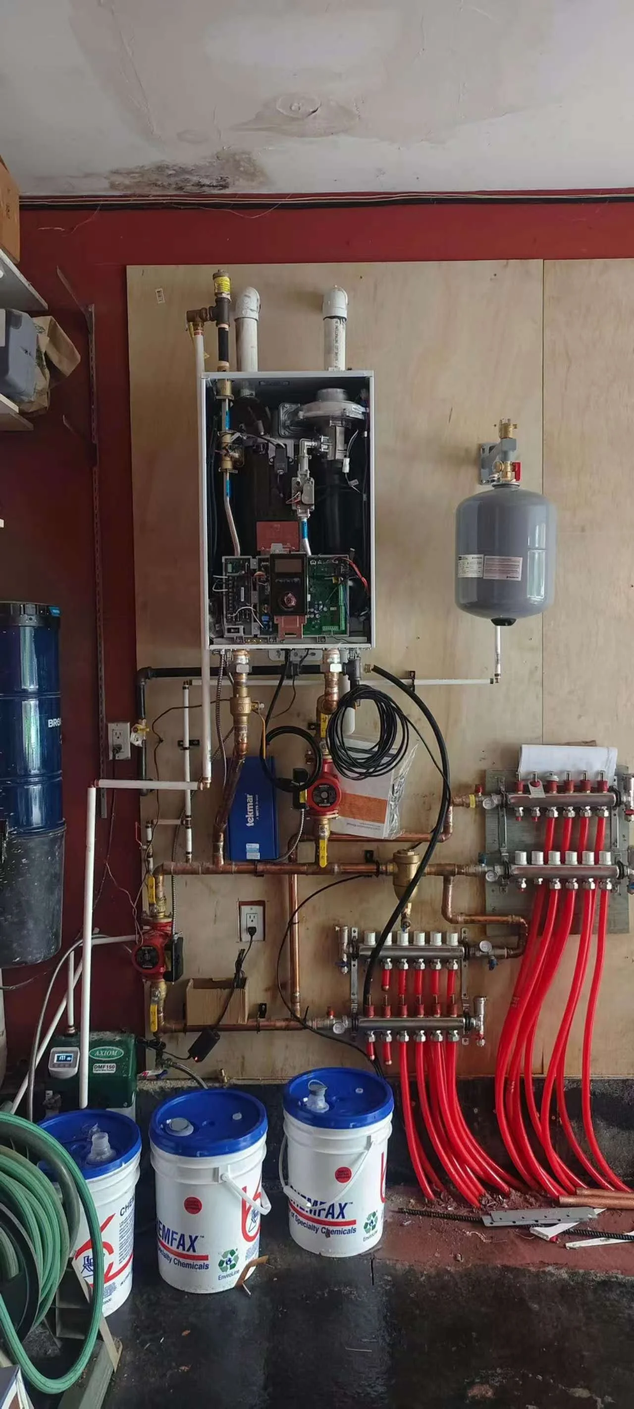A water boiler system with exposed pipes, fittings, and pumps, along with red PEX tubing connected to manifold, and three buckets labeled a chemical supplier, in a utility or maintenance room.