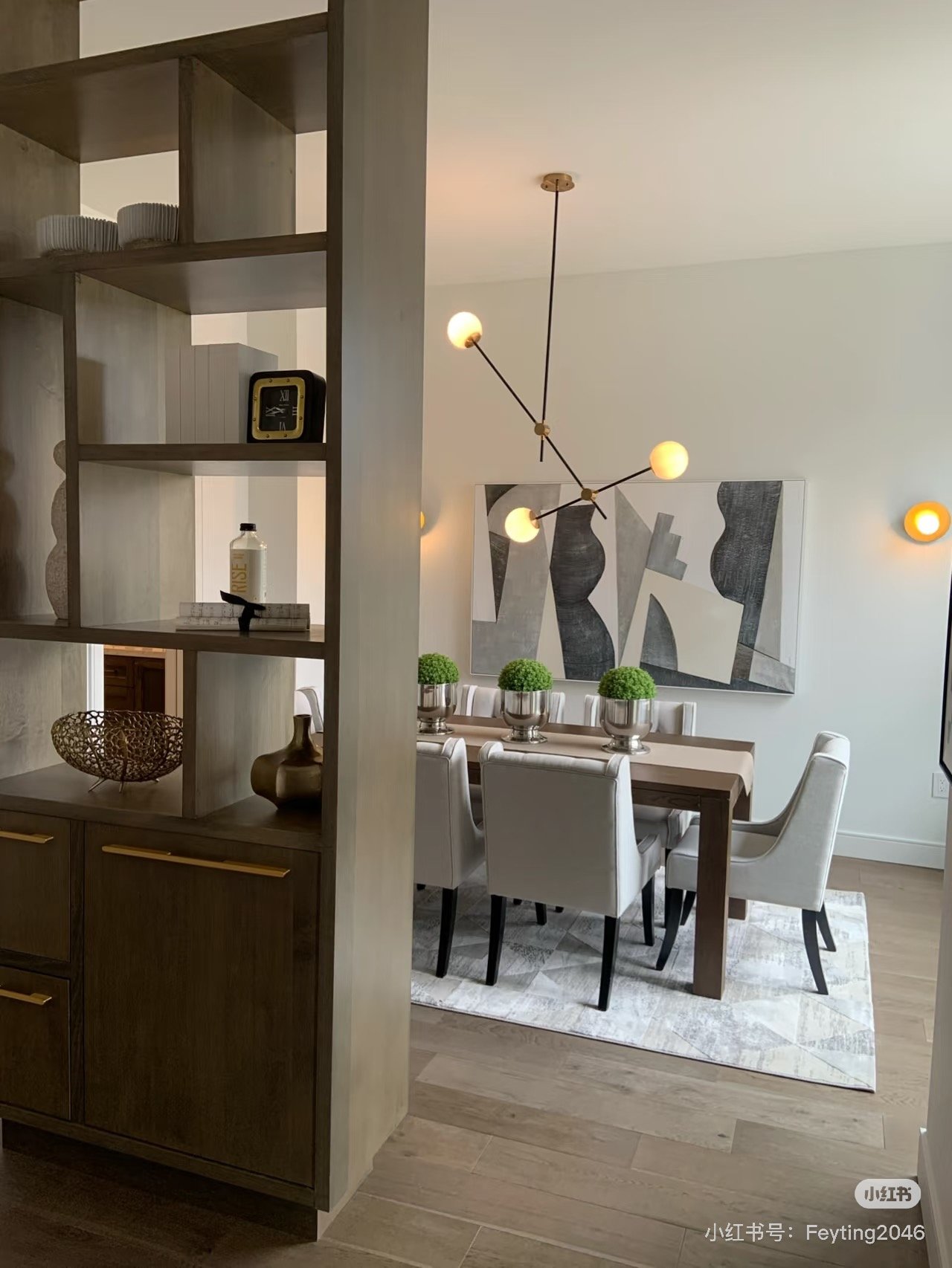 A modern dining room with a wooden dining table surrounded by eight white upholstered chairs, a contemporary abstract painting on the wall, and a geometric chandelier with five spherical bulbs hanging above the table. There are three small green topiary plants in silver pots on the table, and a patterned area rug beneath it.