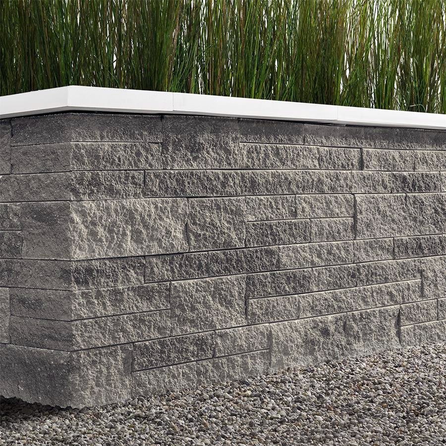 Close-up view of a decorative stone wall with a white top border, set on a gravel ground, with tall green grass behind it.