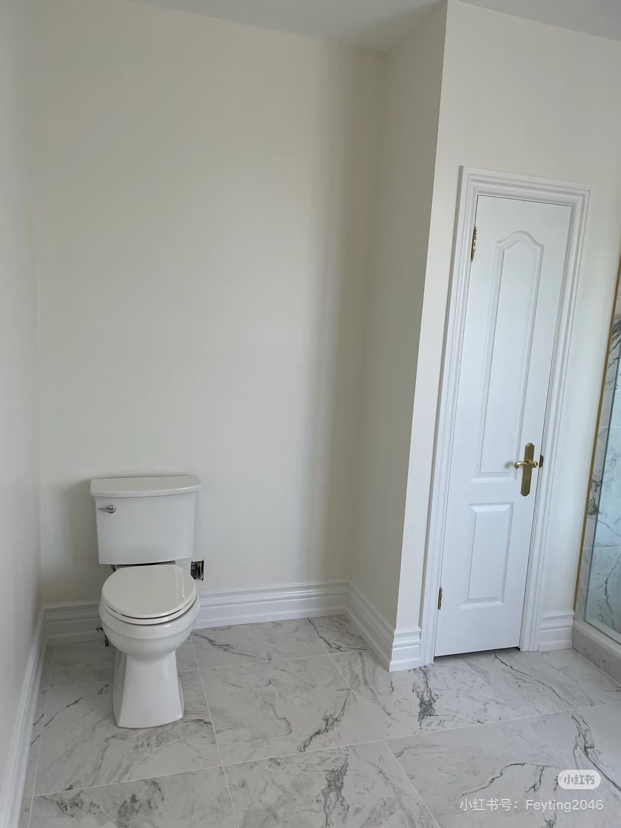 Bathroom with white toilet, marble floor, white paneled door with brass handle, and part of a glass shower enclosure.