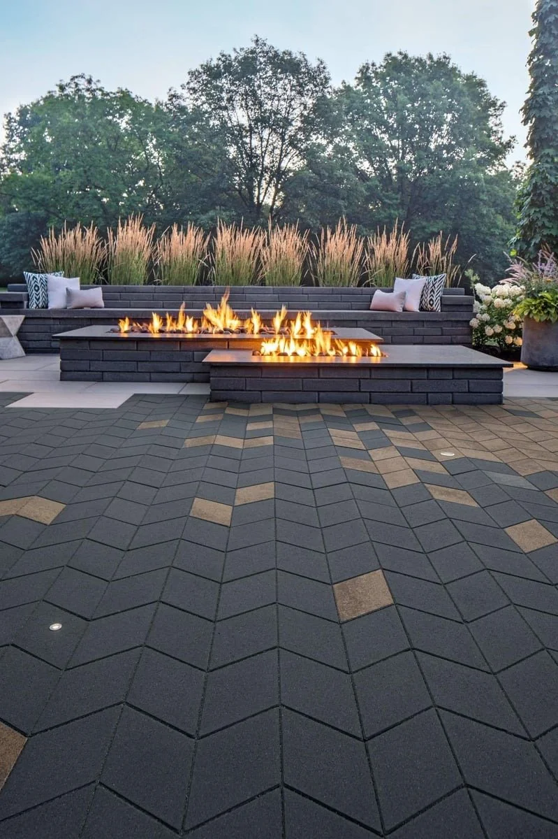 Modern outdoor patio with a fire pit and built-in bench seating, surrounded by decorative pavers and lush greenery.