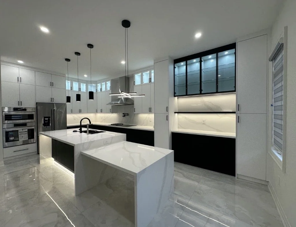 Modern kitchen with white cabinetry, marble countertops, and stainless steel appliances, including a refrigerator and oven, with black accents and pendant lighting.