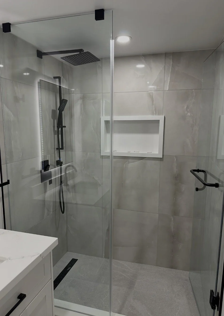 Modern walk-in shower with black fixtures, large gray marble tiles, a ceiling-mounted rainfall showerhead, a handheld showerhead, a built-in wall niche, and clear glass enclosure.