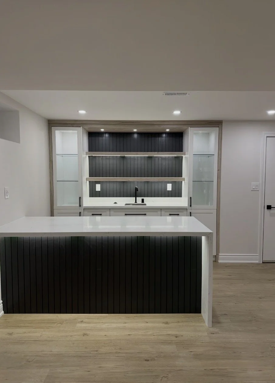 Modern kitchen with white island and black vertical paneling, open shelves, gray background wall, built-in cabinetry, black faucet, and recessed ceiling lights.