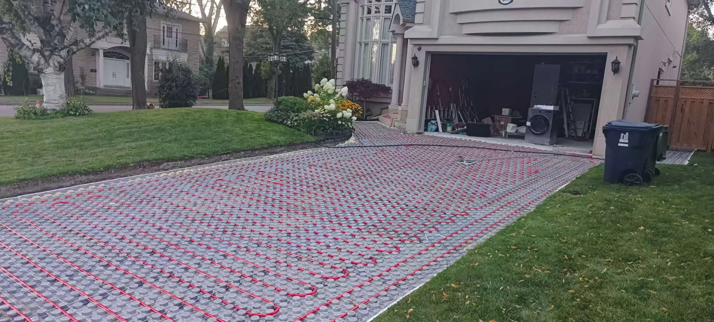 A house driveway is being fitted with underfloor heating with red coils installed over insulation boards, and a garage with various tools and equipment inside. A black trash bin is placed on the side of the driveway.
