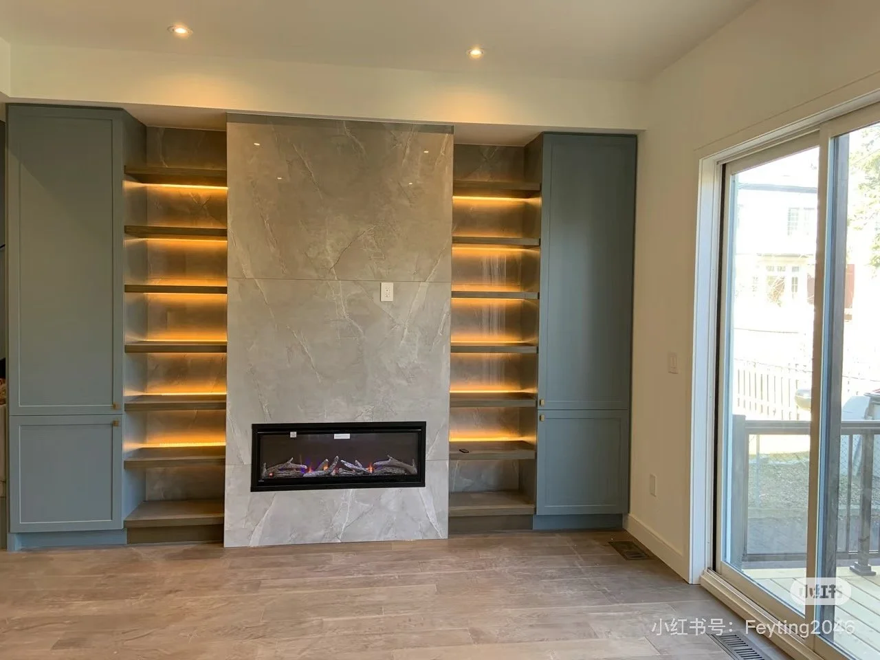 Modern living room with built-in shelves and a fireplace, large sliding glass door leading outside, light-colored walls, and wood flooring.