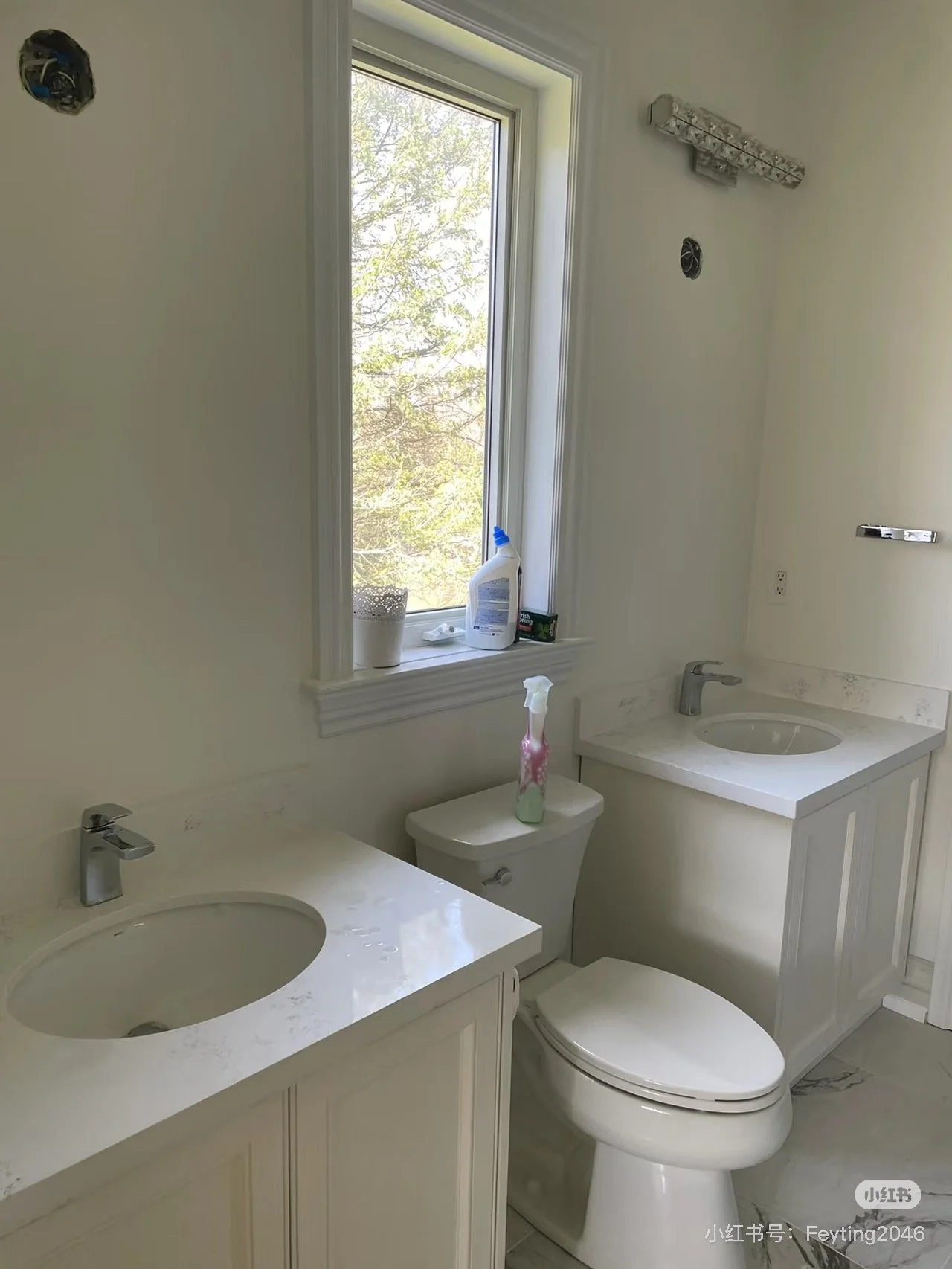 A bathroom with two white sinks and vanity cabinets, a toilet, a window with a view of trees outside, and cleaning supplies on the windowsill.