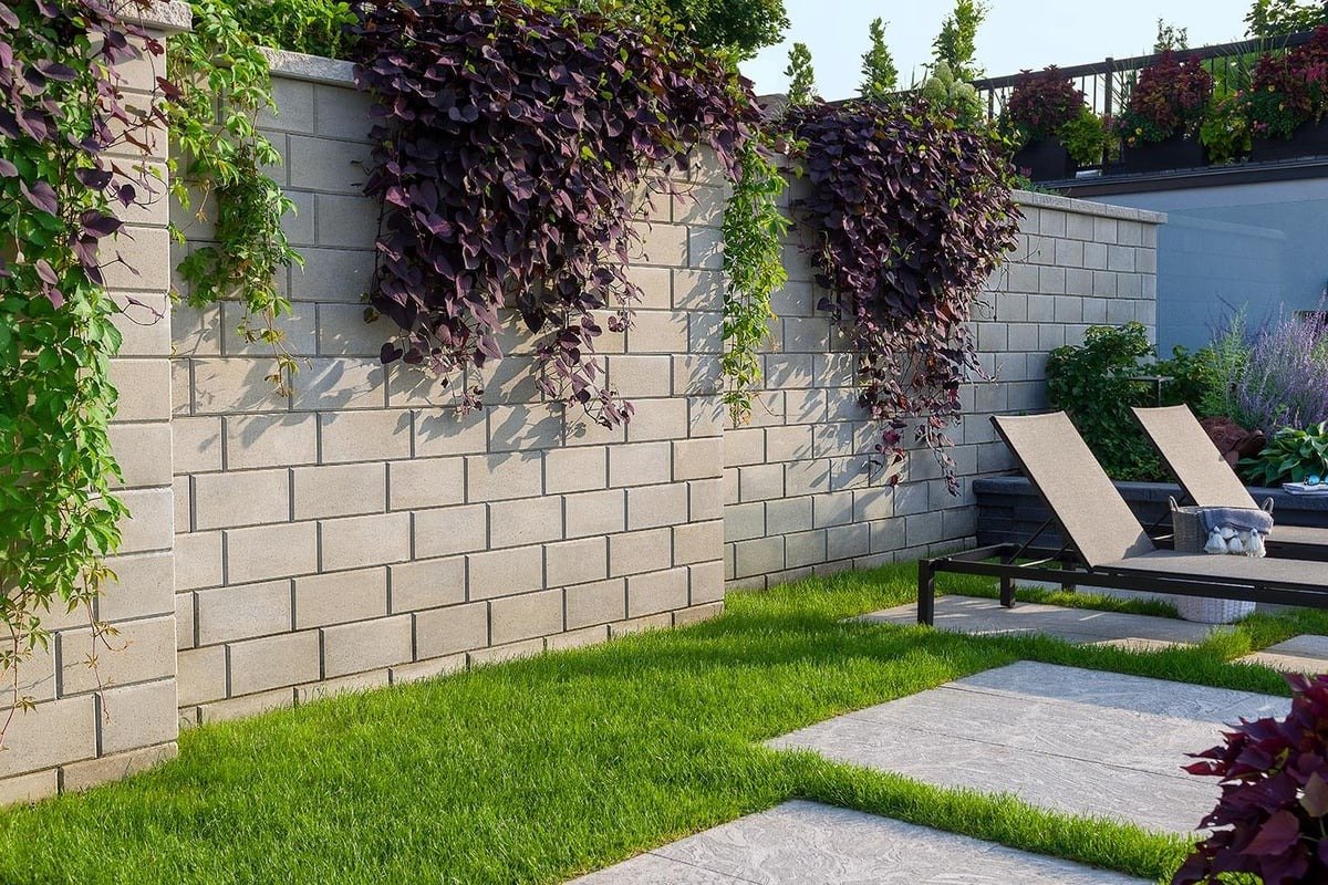 A backyard with a stone brick wall, green grass, purple and green plants, and two lounge chairs with a small table and a basket nearby.