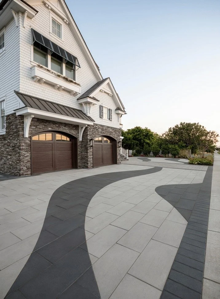 A modern house with a driveway featuring a patterned design of curved dark and light tiles, leading to two garage doors. The house has white siding, stone accents, and black window awnings. Trees and greenery are visible in the background.