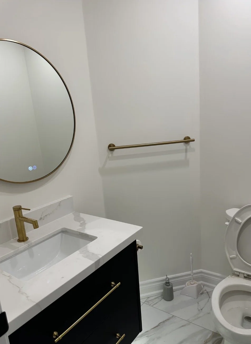 A modern bathroom featuring a round mirror, white marble sink countertop, black vanity with gold hardware, a gold faucet, a towel bar, a toilet with an open lid, and a toilet brush and cleaner nearby.