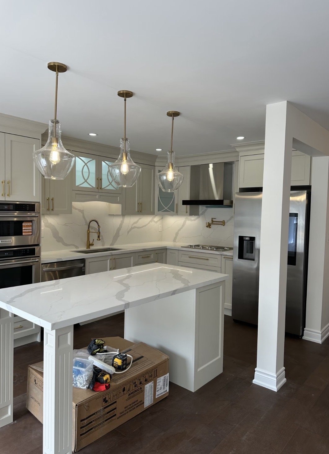 Modern kitchen with white marble countertops, light gray cabinets, gold hardware, stainless steel appliances, hanging glass pendant lights, and a large kitchen island. There are tools and a cardboard box on the island, indicating ongoing installation or remodeling.