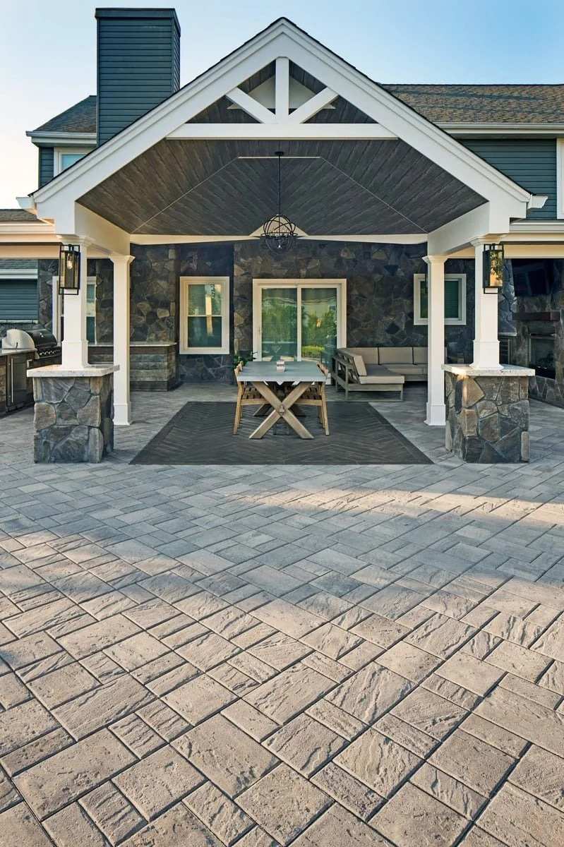 A covered outdoor patio attached to a house with stone and blue siding. The patio features a dining table with benches, a built-in bench with cushions, and a grill. The area is paved with large stone bricks.